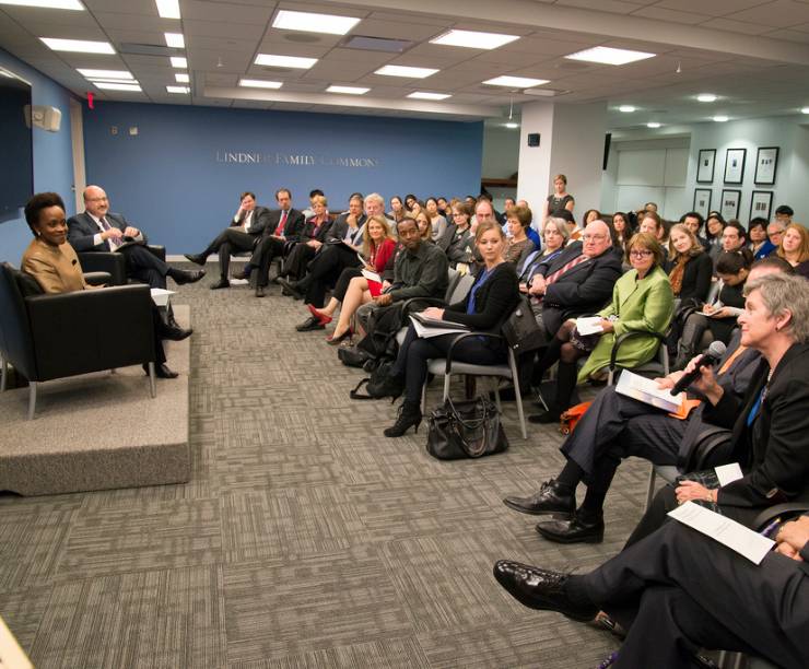 Students sitting in rows of chairs during a lecture. There are two people sitting in the front of the room and one student is asking a question holding a microphone.