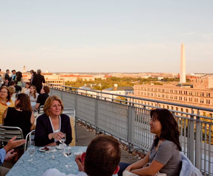 Large gathering of people sitting at outdoor tables, talking, aerial view of city behind them.