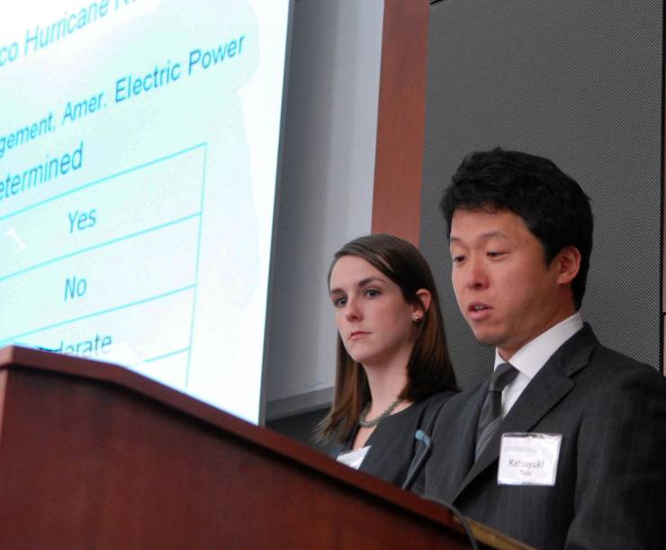 Four students address a lecture hall. Screens behind them show a presentation.