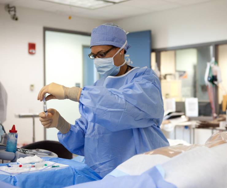 Doctor in scrubs and mask prepares a syringe. A simulation dummy in a drape is on the table in front of her.