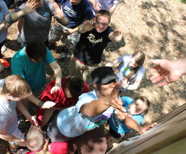 A young man reaches up to a hand extended from above. Other students standing around help lift him up.