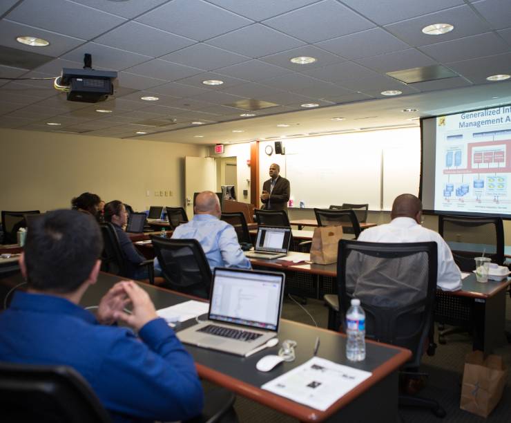 Students on laptops listen to a professor lecture and look at a projection screen.