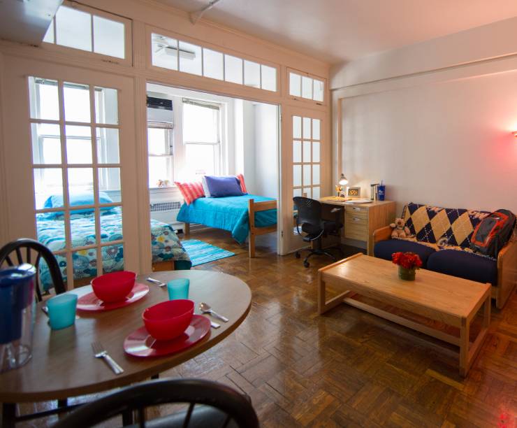 A living room with breakfast and coffee tables, a couch and a desk leading into a two-person bedroom.