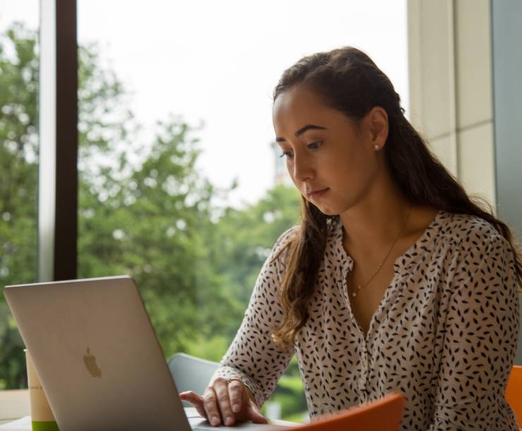 Student sitting in front of a laptop computer in a lounge area with large windows.