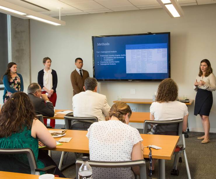 A student speaks at the front of a classroom in front of other students and professors, using a presentation slide on a large television to convey methodology.