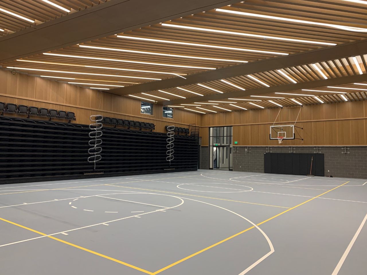 a gymnasium with grey floors, yellow lines, telescoping stadium seating, diagonal ceiling lights, and basketball hoops on each end