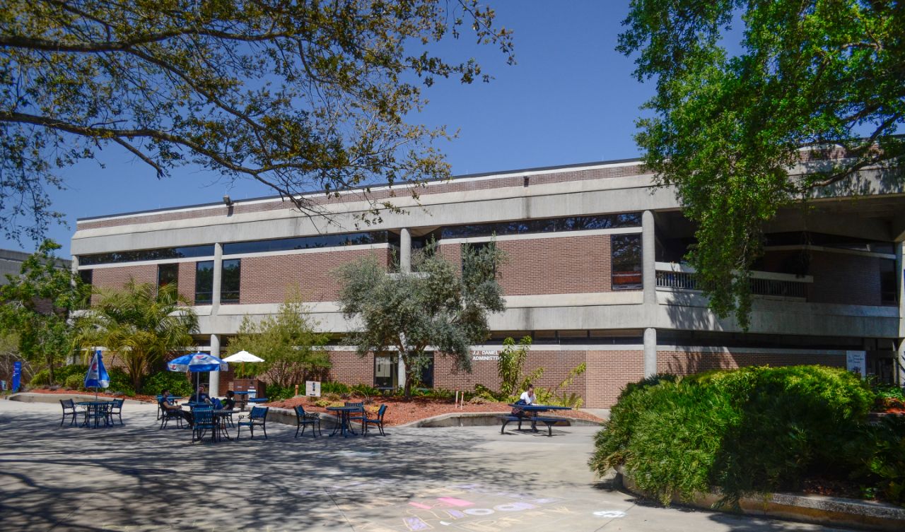 Two-story brick building with white trim sits next to a wide concrete courtyard where students sit at blue tables to study.