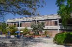 Two-story brick building with white trim sits next to a wide concrete courtyard where students sit at blue tables to study.