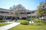 White two-story building with some brick work and covered walkways stretching out from its core sits behind a paved courtyard containing a tall oak tree as well as a bronze sculpture of the globe; In the courtyard are several benches as well as lush landscaping.