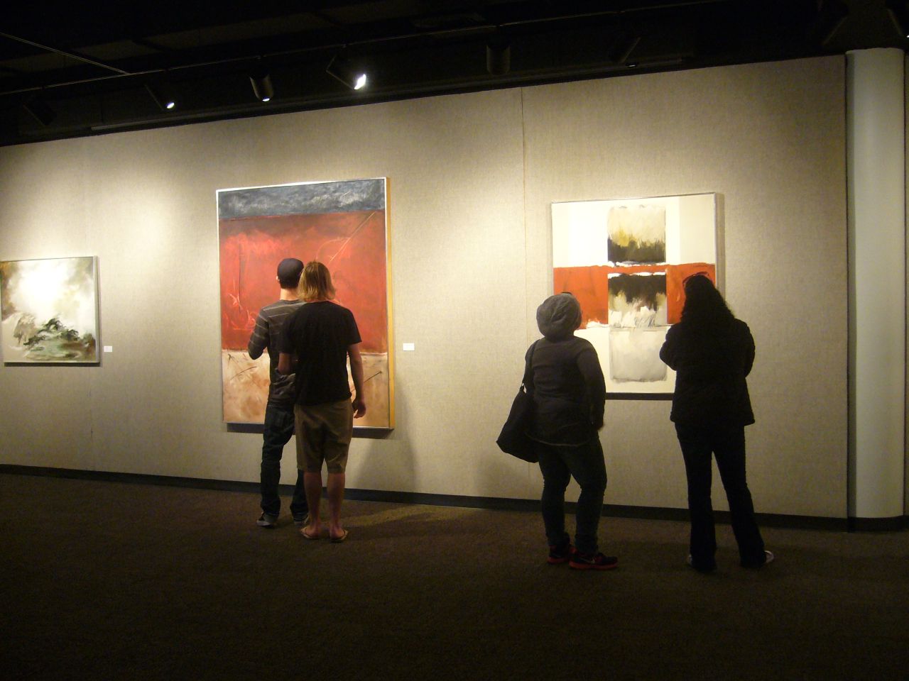 Four students stand gathered around large framed canvases hanging on display in a dark gallery. The canvas to the far left is white and gray; the canvas in the center is blue, red and beige horizontal blocks; and the canvas to the far right is beige and red horizontal blocks with small black and brown squares within each block.