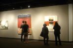 Four students stand gathered around large framed canvases hanging on display in a dark gallery. The canvas to the far left is white and gray; the canvas in the center is blue, red and beige horizontal blocks; and the canvas to the far right is beige and red horizontal blocks with small black and brown squares within each block.