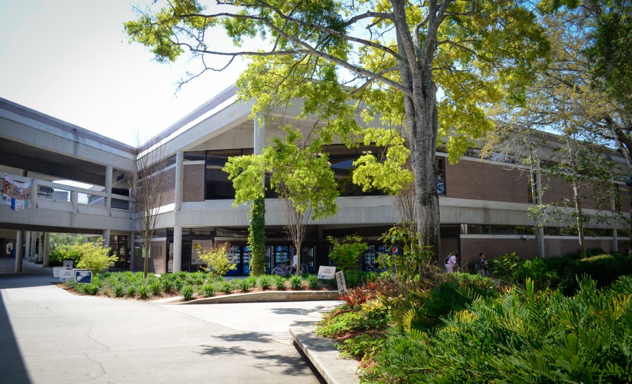 Two-story brown brick building with white trim and a covered walkway extending to the left of the building core sits behind a landscaped plant bed that borders a wide concrete courtyard.