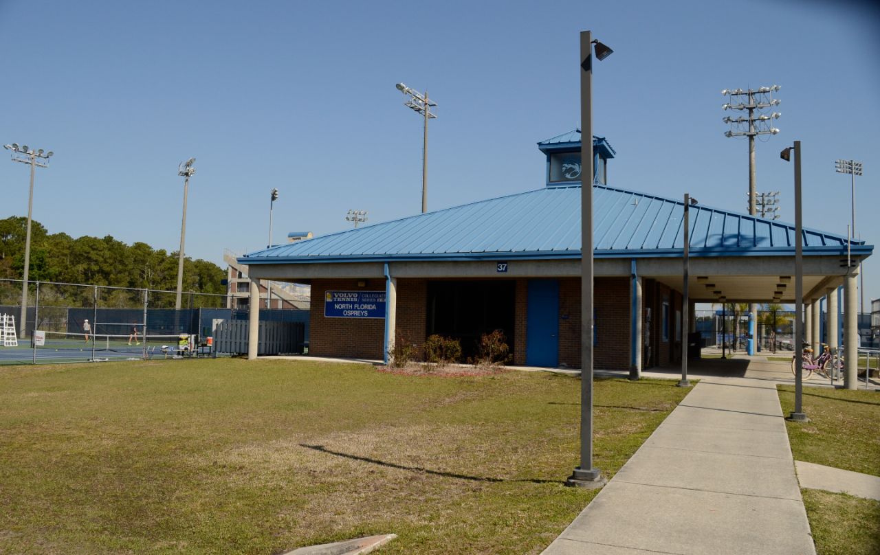 Square one-story brick building with white columns and blue doors and blue metal roof; to the left of the building are tennis courts where student athletes are practicing.