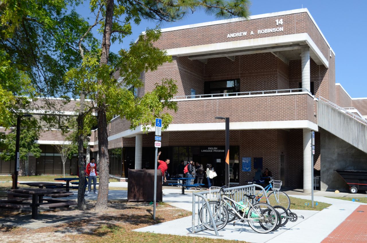 Two-story brick building with white trim has a wide brick balcony on upper floor and a large awning directly underneath at the main corner entrance; in front of the building are bike racks, picnic tables, and a grassy lawn with several pine trees.