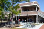 Two-story brick building with white trim has a wide brick balcony on upper floor and a large awning directly underneath at the main corner entrance; in front of the building are bike racks, picnic tables, and a grassy lawn with several pine trees.