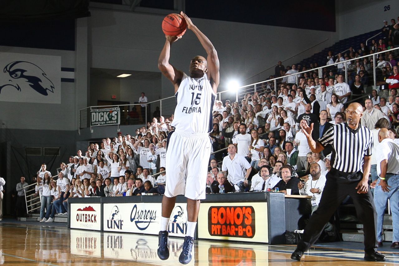 Basketball player wearing white and blue jersey takes a jump shot with basketball in hand on court in front of crowded stands