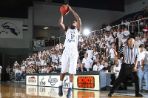 Basketball player wearing white and blue jersey takes a jump shot with basketball in hand on court in front of crowded stands