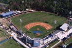 Aerial photo of baseball field with a gray metal and concrete stadium on the home plate corner. Tall stadium lights border the field and dense woods fall on the outside of the center field fence.