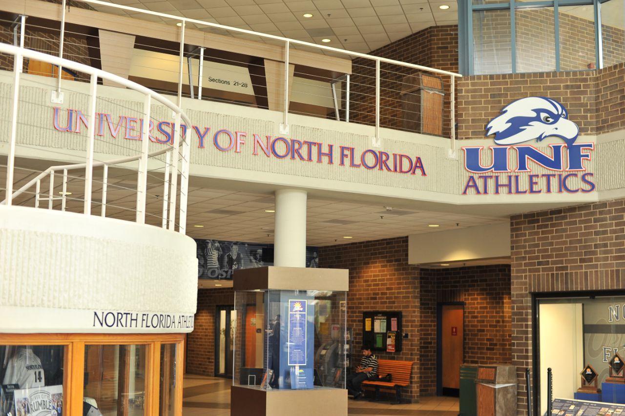 Two-story lobby area with exposed brick walls and white concrete trim; A concrete staircase is at the left and the UNF Athletics logo is hanging from a second floor balcony; A large display case sits in the middle of the lobby and other cases are on the far right wall and underneath the stairs.