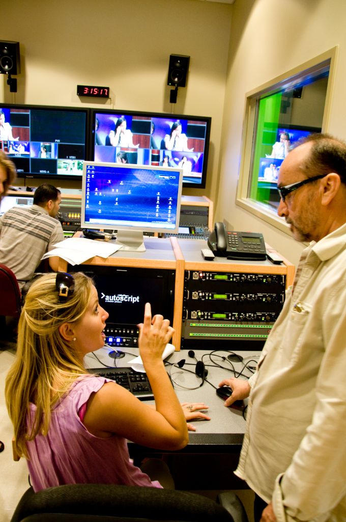 Inside a room with white walls and several desks and large screen TVs, a female student gestures toward a monitor and sound board while consulting with a male professor wearing glasses. The images on the TV screens depict the filming of a news show.