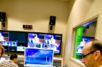 Inside a room with white walls and several desks and large screen TVs, a female student gestures toward a monitor and sound board while consulting with a male professor wearing glasses. The images on the TV screens depict the filming of a news show.