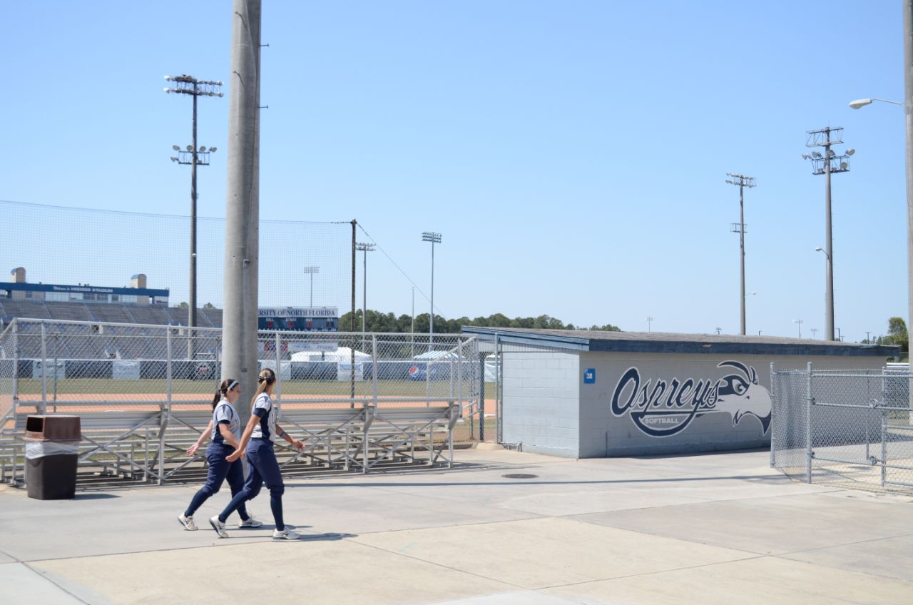 Two female students in blue and gray softball uniforms walk behind the metal bleachers facing a softball field; gray painted concrete dugout is to the right.