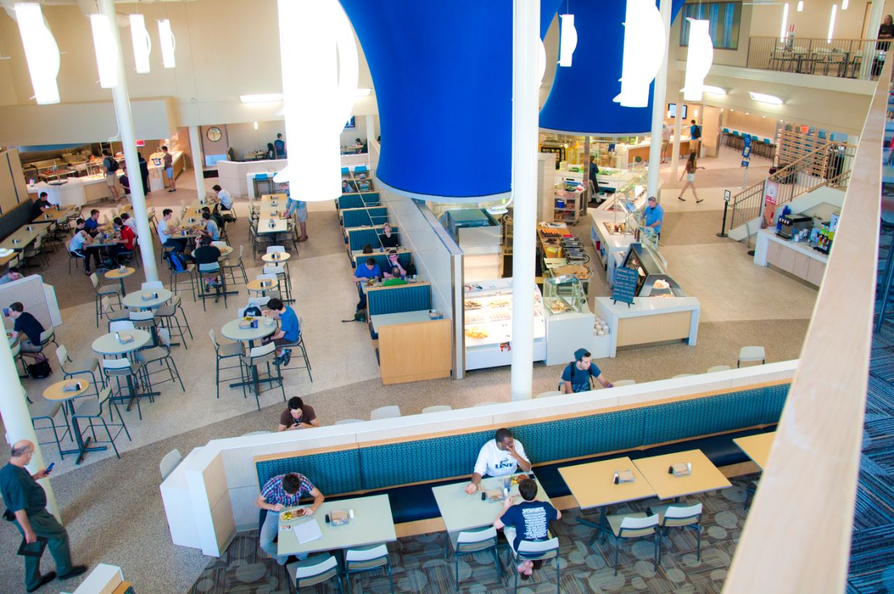 Overhead photo of open floor plan of cafeteria; students are sitting at tables scattered on the near side and left side of the photo; To the right are self-service cafeteria-style food counters with white countertops; Large blue cylinders float above the food stations and thin white cylindrical lighting fixtures hang over the dining areas.