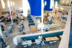 Overhead photo of open floor plan of cafeteria; students are sitting at tables scattered on the near side and left side of the photo; To the right are self-service cafeteria-style food counters with white countertops; Large blue cylinders float above the food stations and thin white cylindrical lighting fixtures hang over the dining areas.