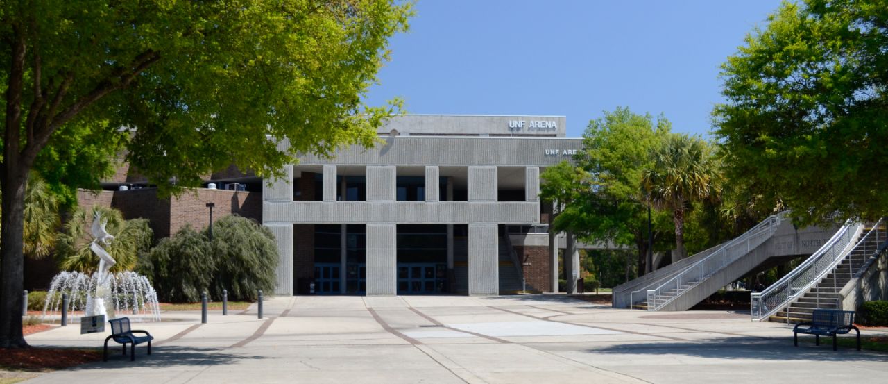 Wide concrete courtyard is bordered by an osprey sculpture and fountain to the left and a sweeping staircase to the right; At the back of the courtyard is a two-story brick building with a large geometric concrete awning