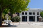 Wide concrete courtyard is bordered by an osprey sculpture and fountain to the left and a sweeping staircase to the right; At the back of the courtyard is a two-story brick building with a large geometric concrete awning