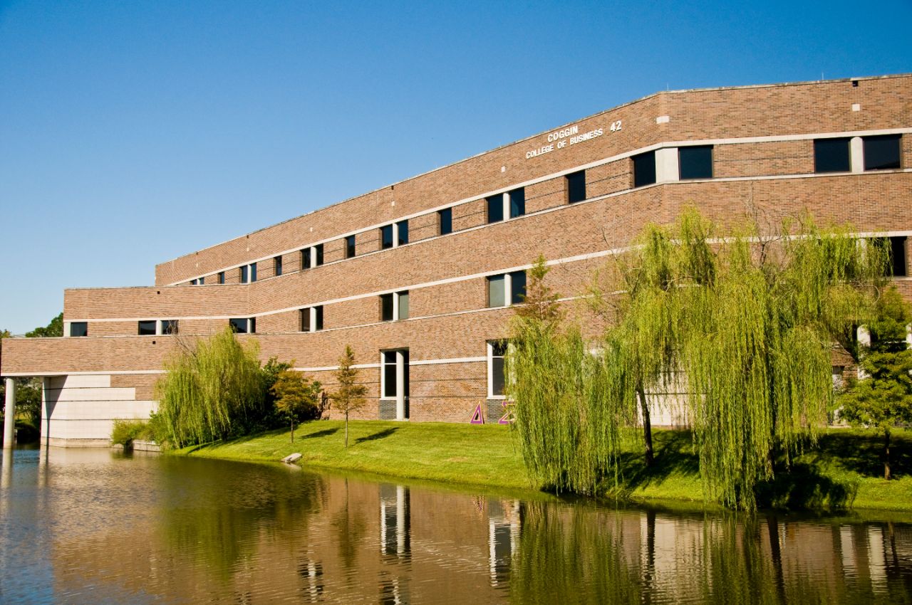 Three-story brick building with square windows sits on the bank of a pond with a grassy shore and a large willow tree.