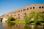 Three-story brick building with square windows sits on the bank of a pond with a grassy shore and a large willow tree.