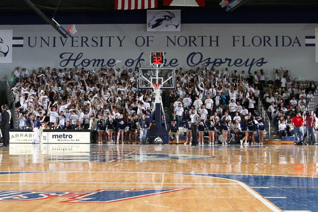 Bleachers are crowded with students doing a cheer with their hands in the air and wearing white UNF t-shirts; Cheerleaders stand in front of stands on sideline of basketball court