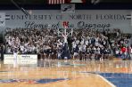 Bleachers are crowded with students doing a cheer with their hands in the air and wearing white UNF t-shirts; Cheerleaders stand in front of stands on sideline of basketball court