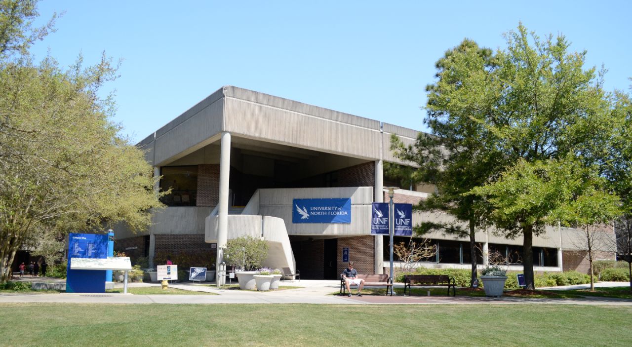 Red brick building with white columns and trim and a tall concrete awning with a white stairwell leading to the second level; in front of the building are potted flowers as well as a pathway that separates the building from a large grassy area. To the left of the building along the path is a wayfinding map; directly in front of the building are two benches and a student is sitting on one of them.