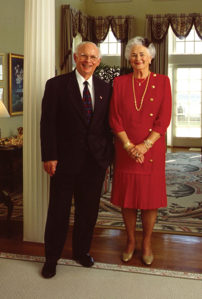 An older couple pose in formal wear in their well-appointed home next to a white column on a hardwood floor between two Oriental rugs with windows behind them and a buffet and paintings to the left of them.