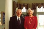 An older couple pose in formal wear in their well-appointed home next to a white column on a hardwood floor between two Oriental rugs with windows behind them and a buffet and paintings to the left of them.