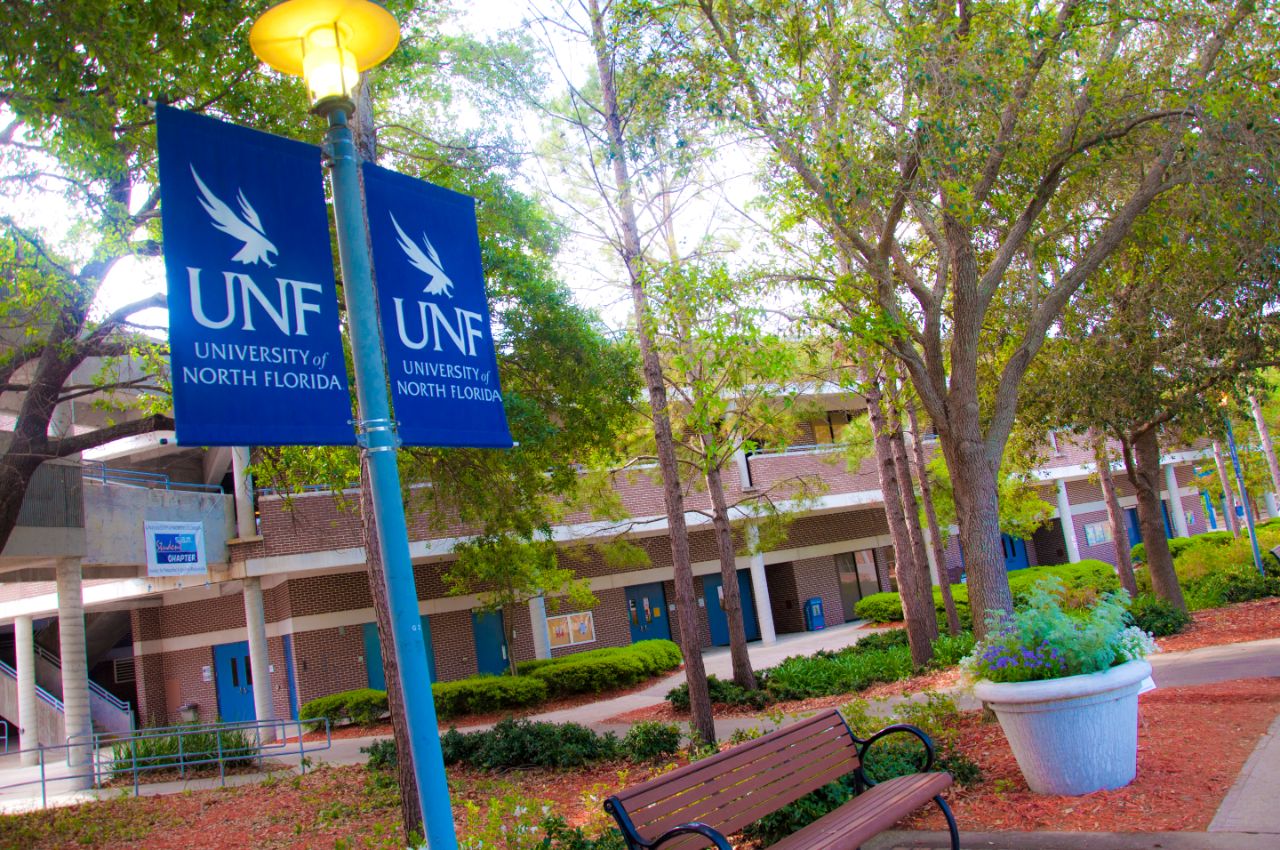 Red brick two-story building with white trim sits along a pathway behind landscaped plant beds where shrubs and trees are growing; in front of the plant beds is a wide concrete sidewalk with a wooden park bench alongside it; To the left of the park bench is a blue lightpole with a blue UNF banner hanging from it.