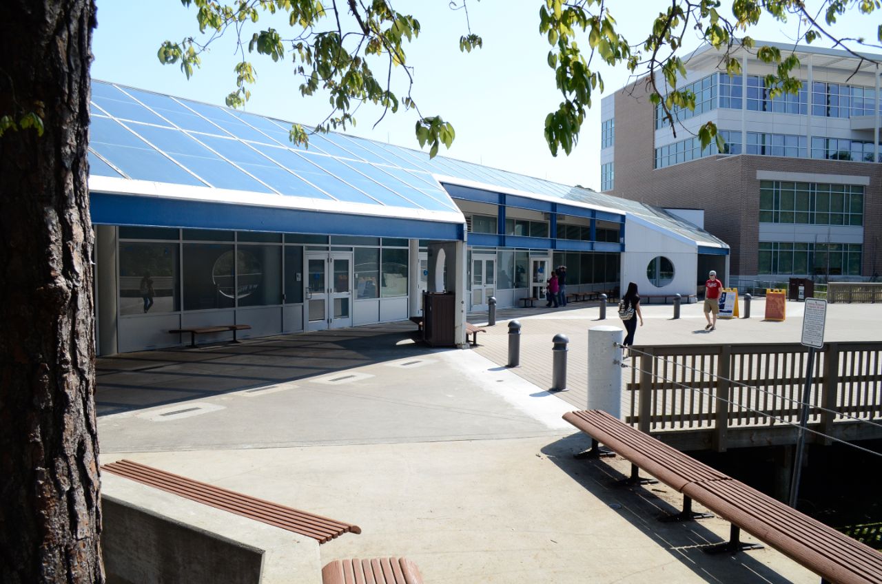 One-story building with large glass windows and a blue awning sits on a wide deck walkway that hangs over a lake; in the background is a three-story brick and glass building; Benches surrounding a tree are located in the foreground