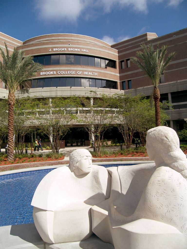 White abstract sculpture of a woman and child in front of blue shallow reflection pool; palm trees are behind the sculpture; beyond the palm trees is a three-story red brick building with white columns and trim.