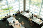 Students study from books and laptop computers in chairs pulled up to two long tables within a corner room with floor-to-ceiling windows overlooking campus