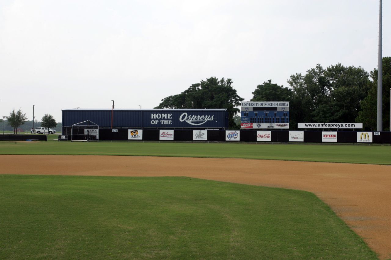 Looking beyond the outfield fence of a baseball field, a one-story blue and white metal building extends between the left field line and a scoreboard in centerfield. The building has the words “Home of the Ospreys” painted on the outside.