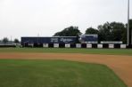 Looking beyond the outfield fence of a baseball field, a one-story blue and white metal building extends between the left field line and a scoreboard in centerfield. The building has the words “Home of the Ospreys” painted on the outside.