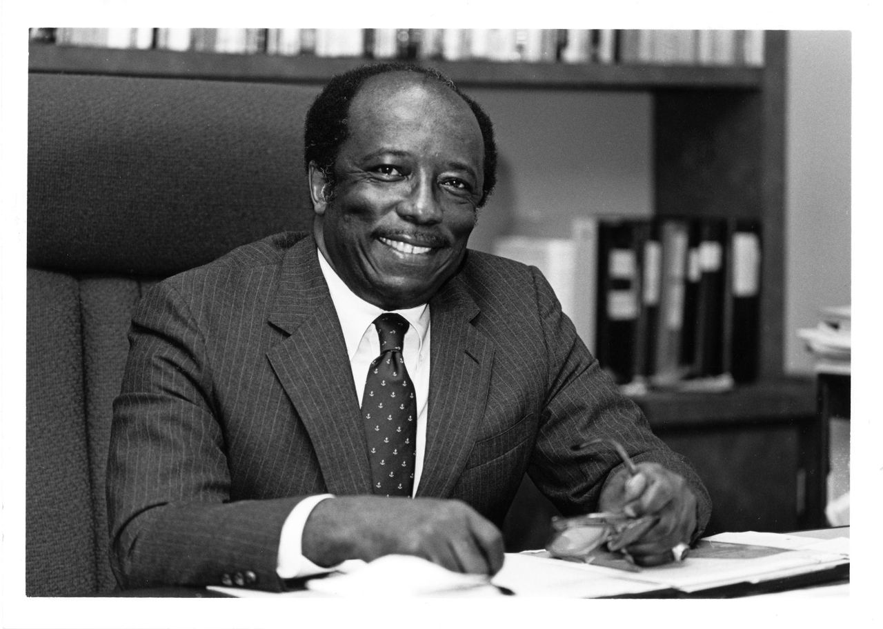 An African American male seated behind a desk wearing a suit and holding a pair of glasses in his hand, smiles for the camera, while taking a break from working with papers on his desk.