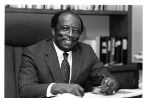 Andrew RobinsonAn African American male seated behind a desk wearing a suit and holding a pair of glasses in his hand, smiles for the camera, while taking a break from working with papers on his desk.