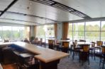 Room with floor-to-ceiling windows and blue patterned carpet with a bar on the left side that has refrigerators beneath a beige countertop and high top tables and chairs across the window area to the right.