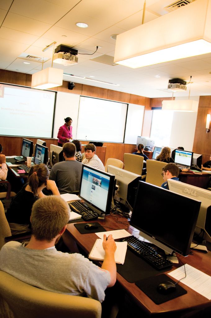 Students sit at rows of desks with computers at each seat; students are watching a professor in a pink suit give a lecture in front of a projector screen in a classroom with wood-paneled walls.