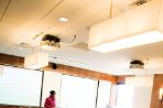 Students sit at rows of desks with computers at each seat; students are watching a professor in a pink suit give a lecture in front of a projector screen in a classroom with wood-paneled walls.