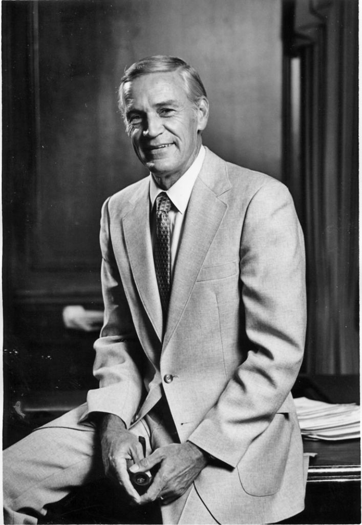 A black and white photograph of an older Caucasian male sitting on the edge of a 1960s-style desk, smiling for the camera.