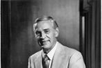 A black and white photograph of an older Caucasian male sitting on the edge of a 1960s-style desk, smiling for the camera.